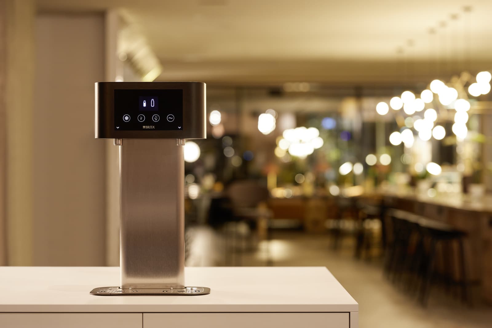 Wasserspender in der Gastronomie: BRITA Zapfstelle mit Bedienfeld auf einer hellen Theke, dahinter unscharf der abendlich beleuchtete Gastraum eines Restaurants.