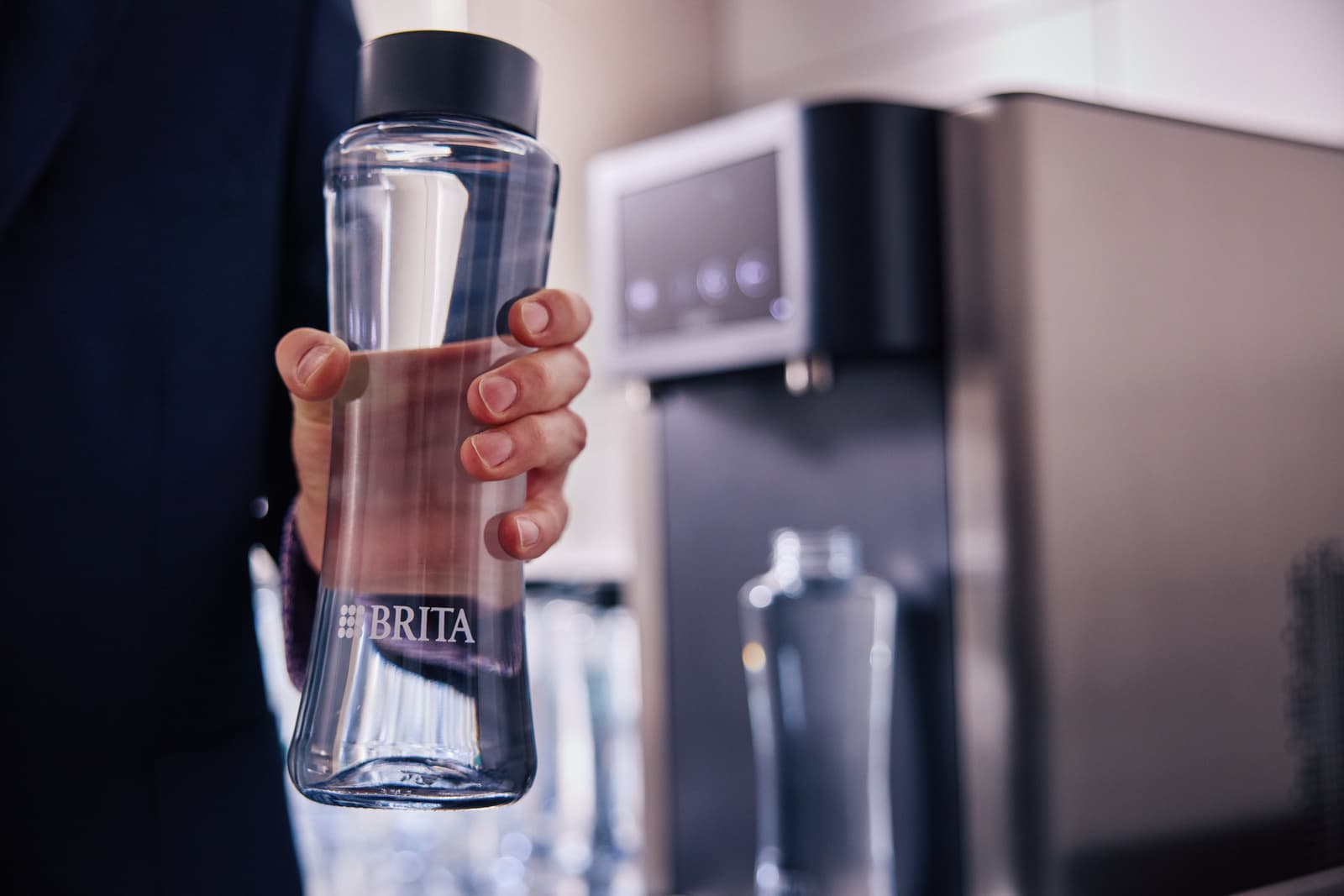 Eine Hand hält eine frisch gefüllte BRITA Trinkflasche, im unscharfen Hintergrund der Wasserspender und weitere bereitstehende Flaschen.