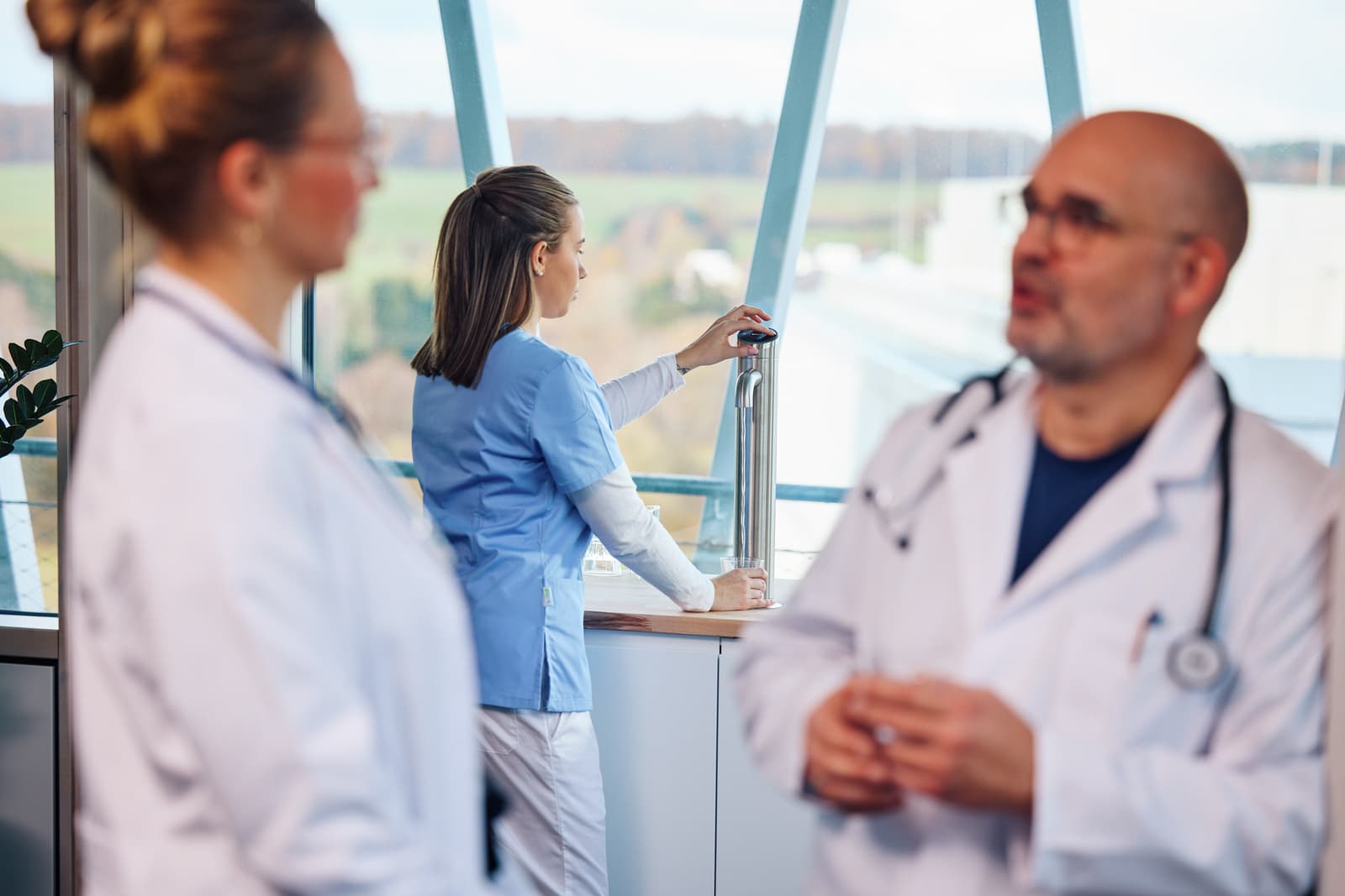 Wasserspender in der Klinik: Eine Pflegekraft in blauer Dienstkleidung füllt am Edelstahl-Auslass einer Fensterbanktheke ein Glas, im unscharfen Vordergrund zwei Personen in weißen Kitteln.