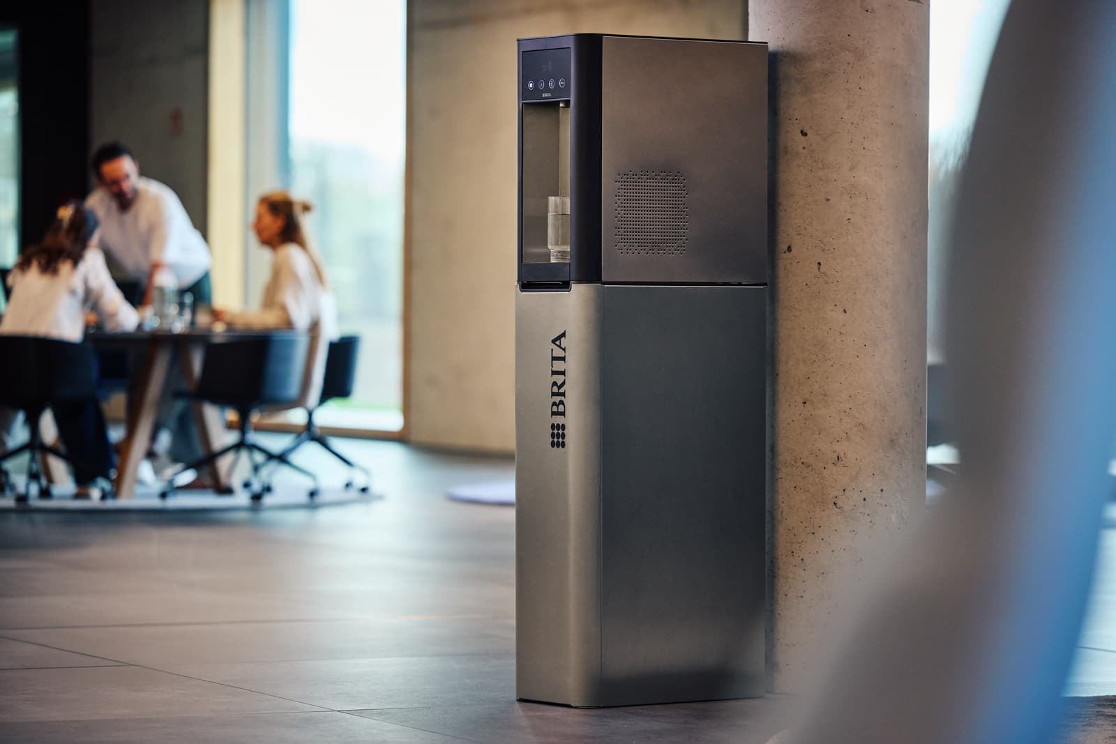 Wasserspender als Standgerät im Flur: BRITA Spender mit Entnahmenische an einer Betonstütze, im unscharfen Hintergrund eine Gruppe an einem Tisch.