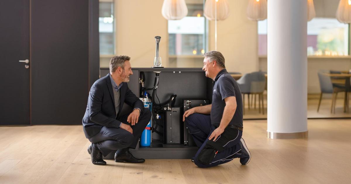 Servicetechniker und Kunde knien vor dem geöffneten Unterschrank eines BRITA Extra C-Tap: Filter, Technik und CO₂-Zylinder liegen frei, die Zapfstelle sitzt auf der Theke darüber.