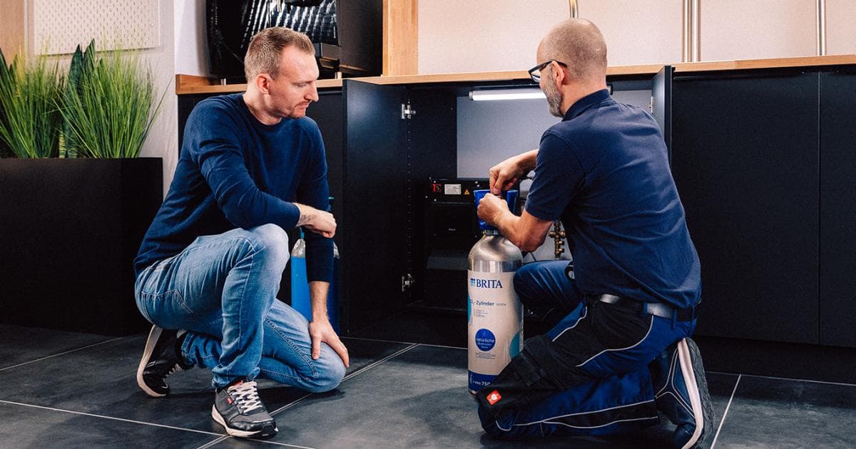 Techniker und Kunde vor dem geöffneten Unterschrank eines BRITA Wasserspenders beim Wechsel des CO₂-Zylinders.