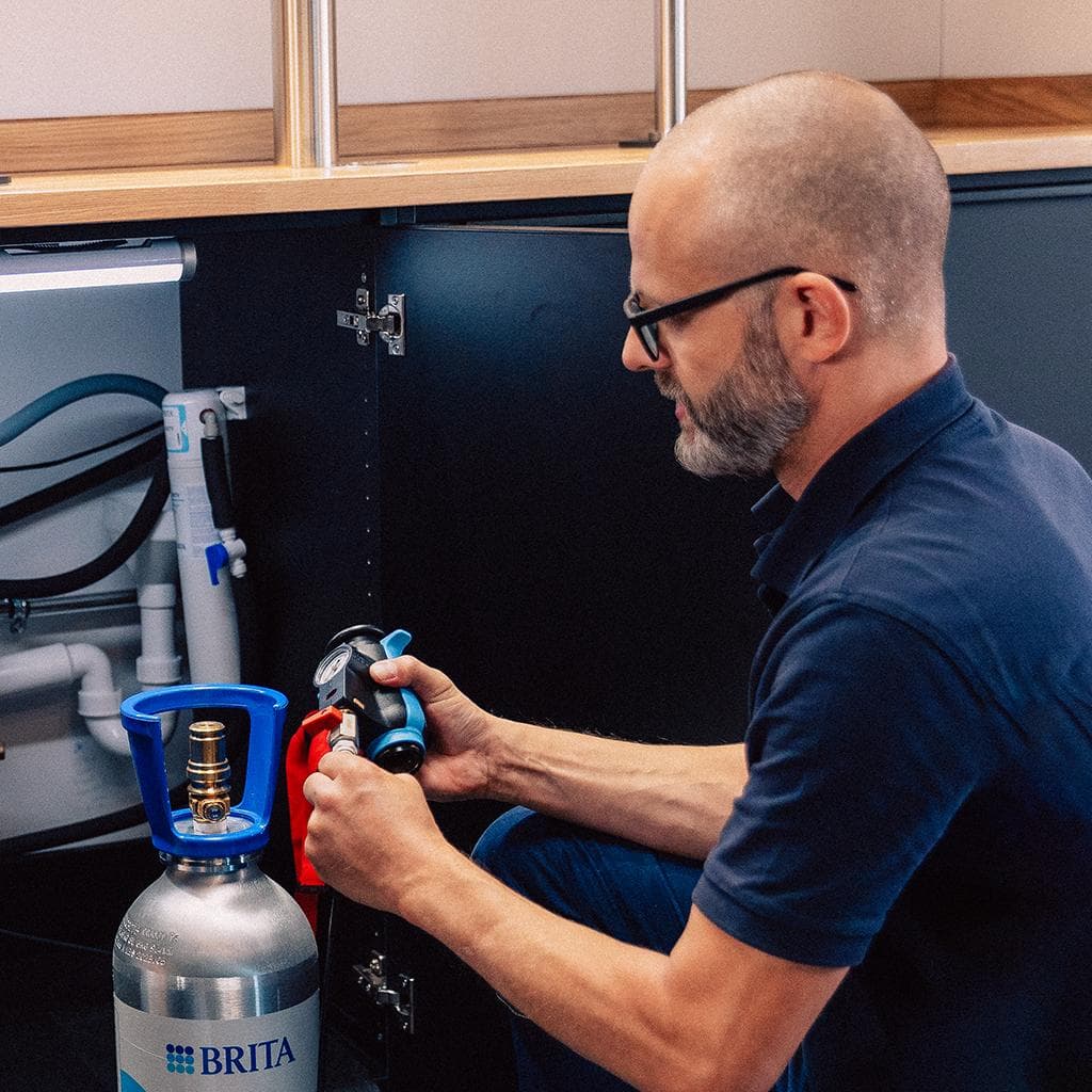 Techniker bei der Wartung eines BRITA Wasserspenders: Er schließt im geöffneten Unterschrank den CO₂-Zylinder an.
